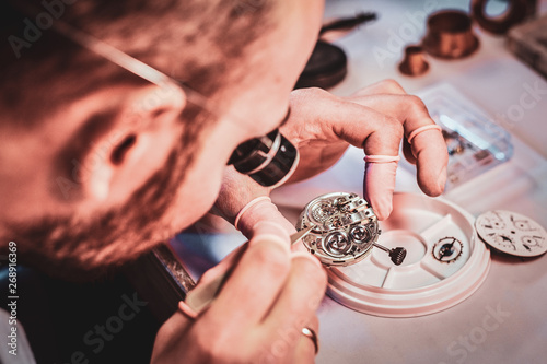 Mature clockmaster is fixing old watch for a customer at his busy repairing workshop.