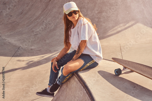Beautiful young woman in sunglacces and cap is sitting at skatepark on the ramp with her longboard.  photo
