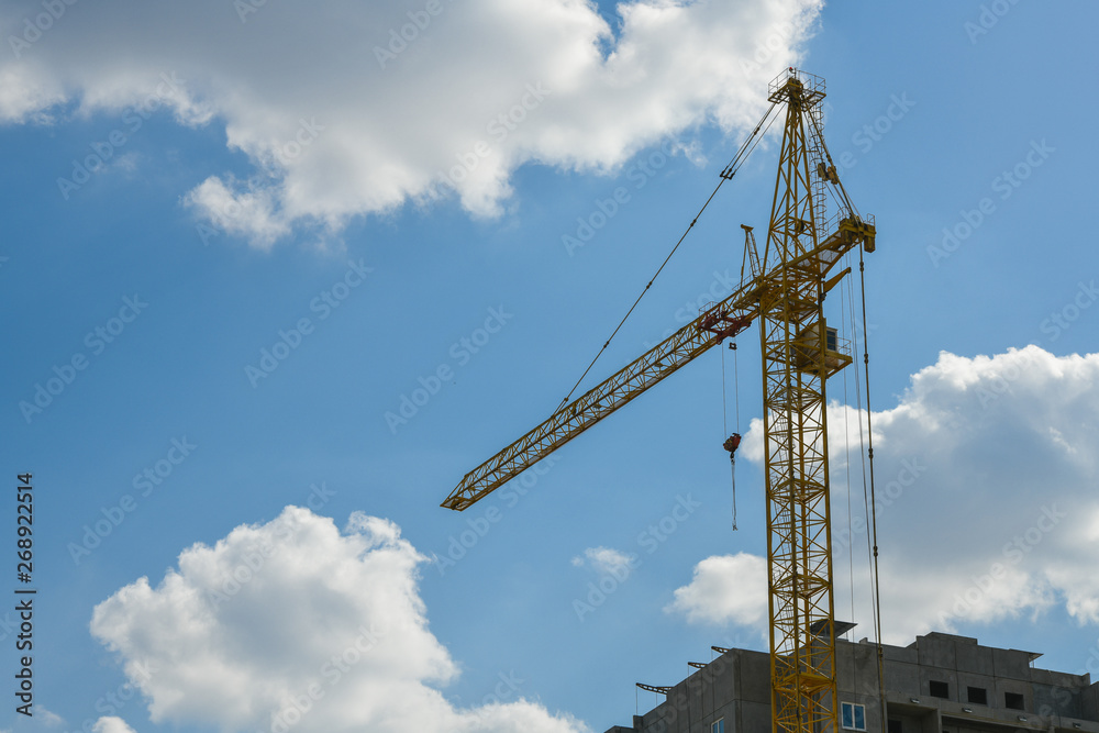 high-rise construction crane with a long arrow of yellow color against the blue sky over a new multi-storey building of concrete and brick under construction