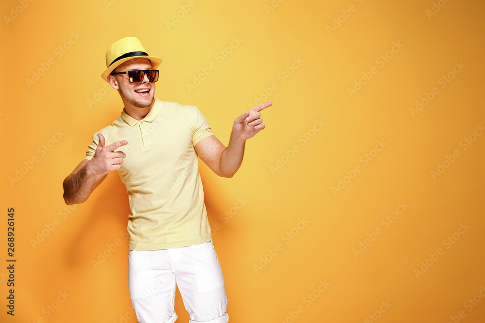 Positive young male in trendy outfit smiling and pointing away while standing against yellow background