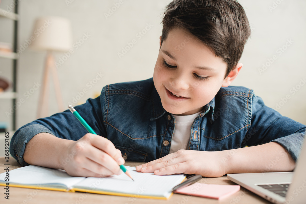 cheerful boy writing in notebook while sitting at desk and doing homework cheerful boy writing in notebook while sitting at desk and doing homework