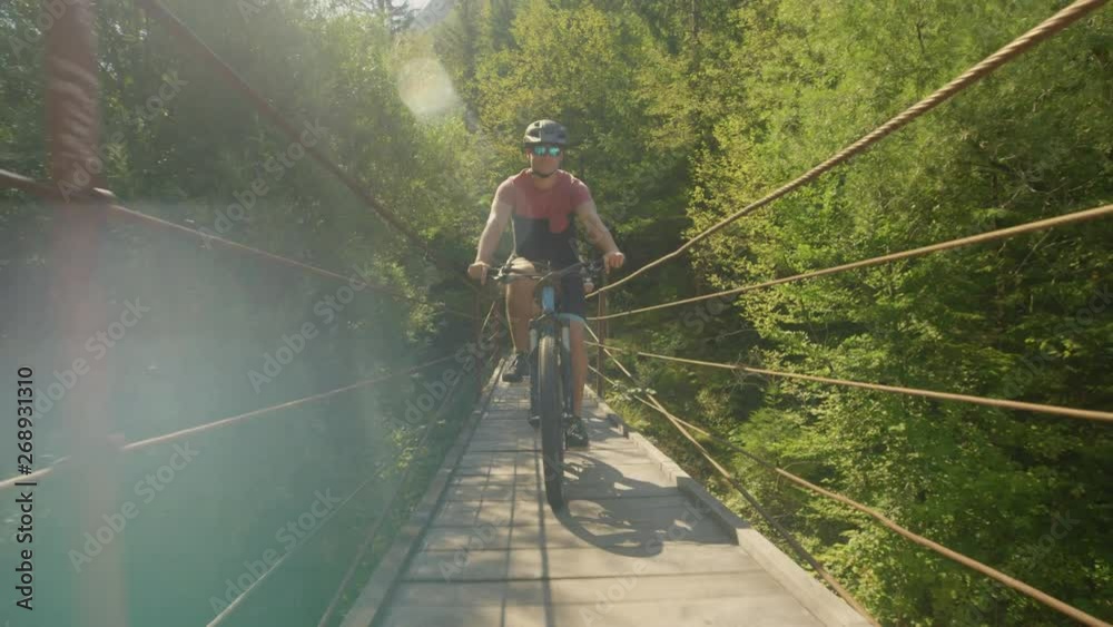 LENS FLARE: Cheerful Caucasian man rides a mountain bike across a suspension bridge on sunny day. Two young friends exploring the Soca river valley cross a bridge while riding electric bicycles. LENS FLARE: Cheerful Caucasian man rides a mountain bike across a suspension bridge on sunny day. Two young friends exploring the Soca river valley cross a bridge while riding electric bicycles.