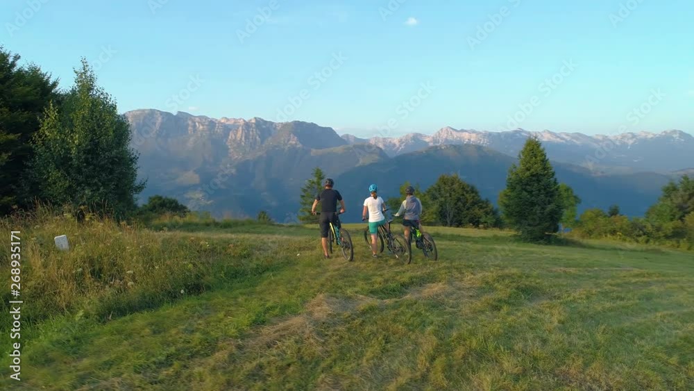 DRONE: Three mountain bikers stop and observe the picturesque morning landscape before exploring the Slovenian countryside. Young tourists riding ebikes stop to look around the scenic evening nature.