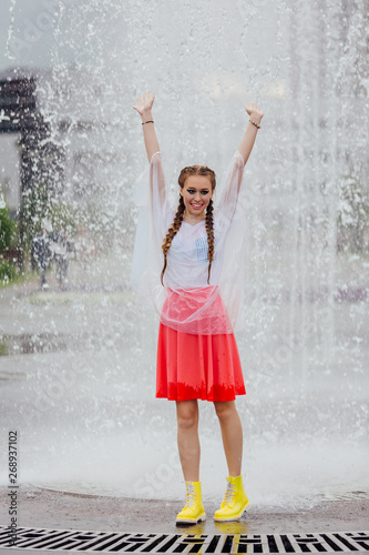 Young wet pretty girl with two braids in yellow boots and with transparent umbrella stands inside of fountain.