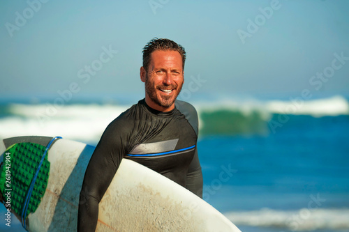 handsome and attractive surfer man in neoprene swimsuit carrying surf board smiling happy and cheerful after surfing enjoying Summer water sport and holidays