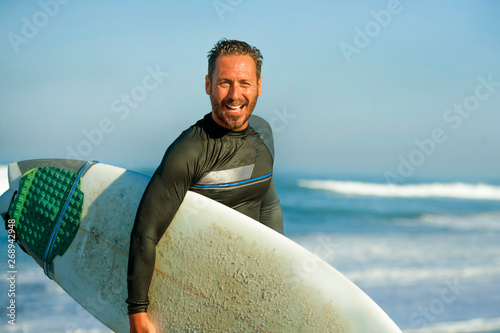 handsome and attractive surfer man in neoprene swimsuit carrying surf board smiling happy and cheerful after surfing enjoying Summer water sport and holidays