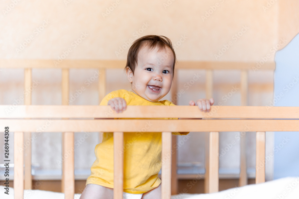 Happy baby girl with dark hair laughing and smiling in yellow t-shirt mixed race kazakh and german standing in cot crib Happy baby girl with dark hair laughing and smiling in yellow t-shirt mixed race kazakh and german standing in cot crib
