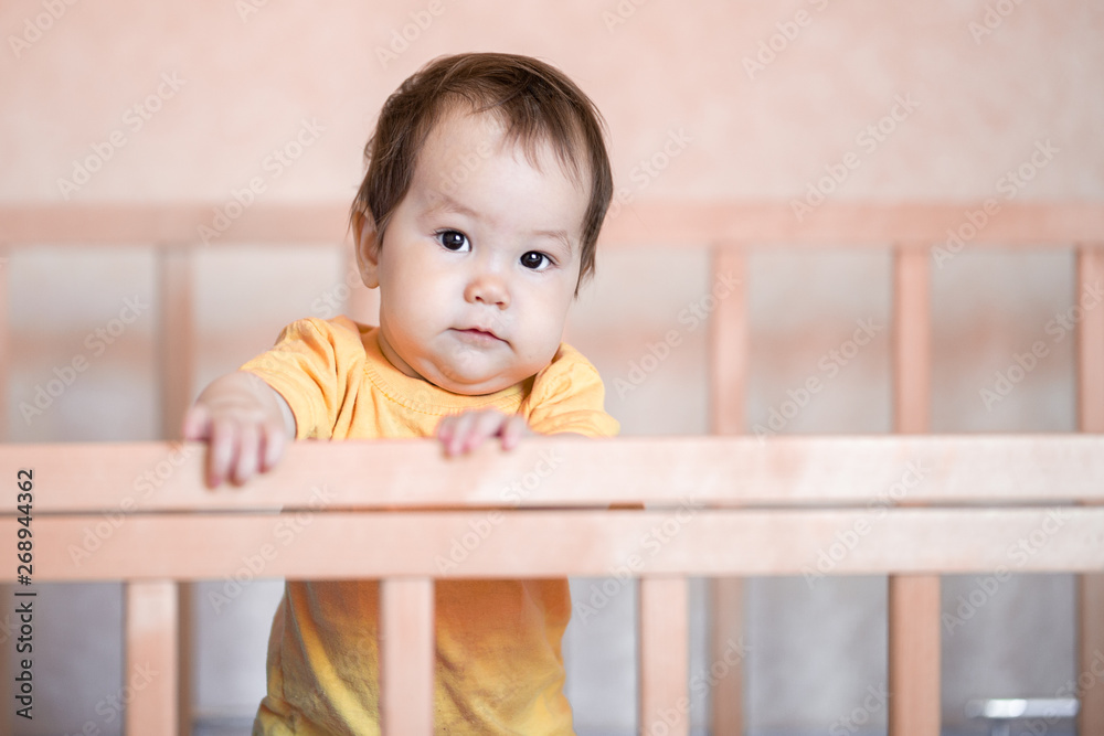 Cute little baby girl with dark hair standing in crib at home in yellow t-shirt Cute little baby girl with dark hair standing in crib at home in yellow t-shirt