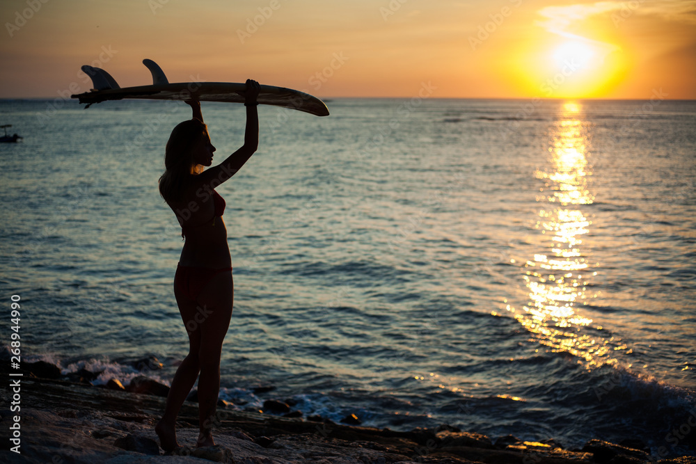 silhouette of a girl in backlight holding a surfboard above her head. Sunset on the sea coast