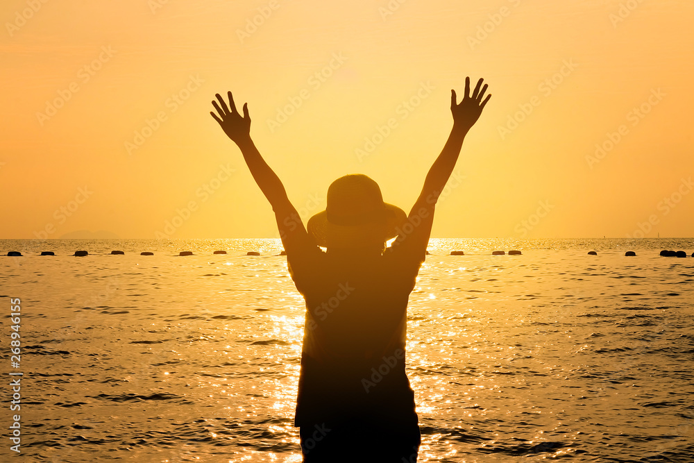 The silhouette of the happy woman holds arms up while on the seaside at the evening sunset