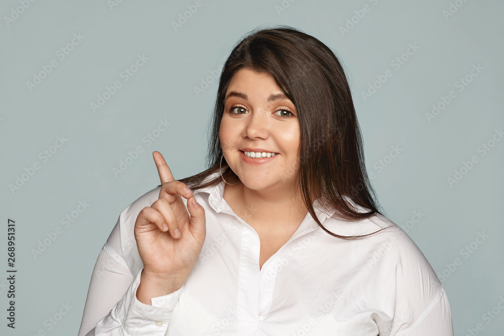 Gorgeous joyful young plus size businesswoman in good mood posing isolated at gray wall, looking at camera with happy confident smile, showing something to you, pointing index finger sideways Gorgeous joyful young plus size businesswoman in good mood posing isolated at gray wall, looking at camera with happy confident smile, showing something to you, pointing index finger sideways