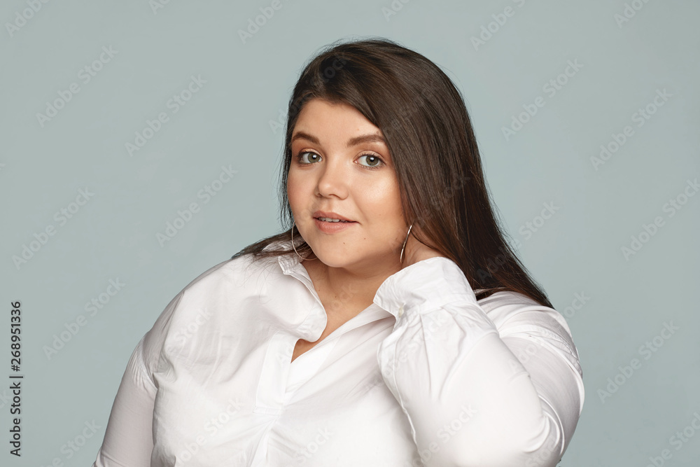 Picture of stylish young overweight female employee wearing white shirt and large round earrings touching her neck. Neat beautiful chubby woman posing against gray scopyspace tudio wall background Picture of stylish young overweight female employee wearing white shirt and large round earrings touching her neck. Neat beautiful chubby woman posing against gray scopyspace tudio wall background