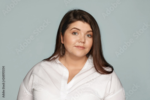 Horizontal studio portrait of adorable beautiful young overweight European woman with loose black hair and chubby cheeks wearing round earrings and xxl white shirt posing at blank wall, smiling