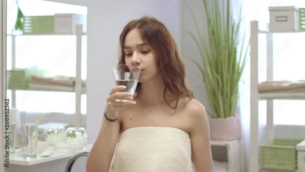 Woman wrapped in towel taking capsule tablet and drinking water in bathroom Woman wrapped in towel taking capsule tablet and drinking water in bathroom