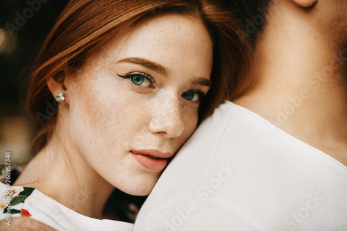 Fotografie Close up portrait of a amazing red haired woman with freckles and green eyes looking at camera while leaning her head on her boyfriend back outside while dating