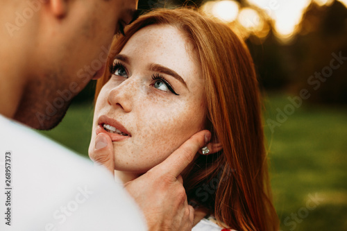 Portrait of a red haired woman with freckles and green eyes looking at her boyfriend while he is touching her lips with a finger against sunset while dating