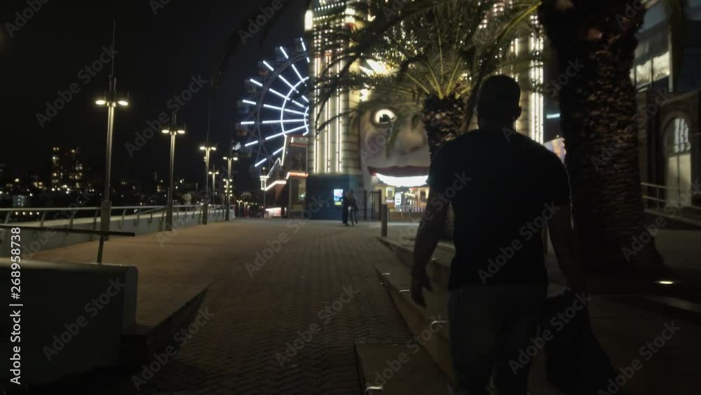 Camera Smoothly Follows Man Walking Towards Theme Park at Night Camera Smoothly Follows Man Walking Towards Theme Park at Night