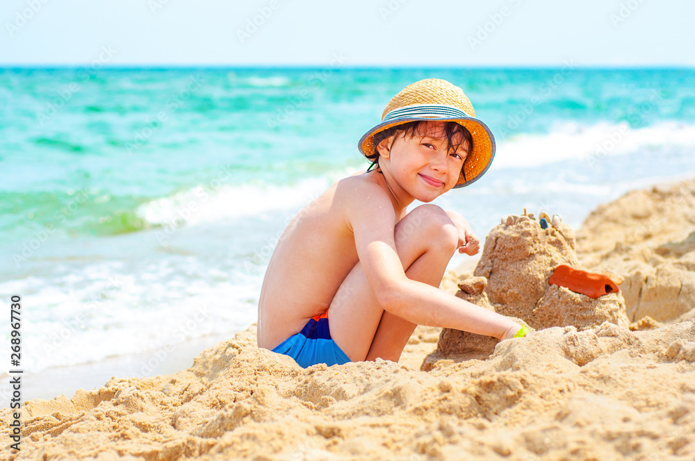 A happy boy is building a sand castle on the shore of a turquoise sea on a sunny day. Children's and family rest on the sea in the summer. - obrazy, fototapety, plakaty 