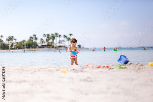 Cute little curly baby play with water and sand on beach at the seaside. Enjoying a lovely summer day. Vacation concept © olenap