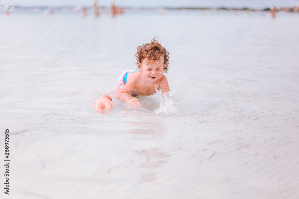 Cute little curly baby play with water and sand on beach at the seaside. Enjoying a lovely summer day. Vacation concept