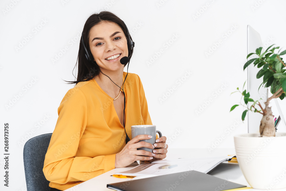 Photo of happy woman wearing microphone headset drinking cup of tea while working in office Photo of happy woman wearing microphone headset drinking cup of tea while working in office