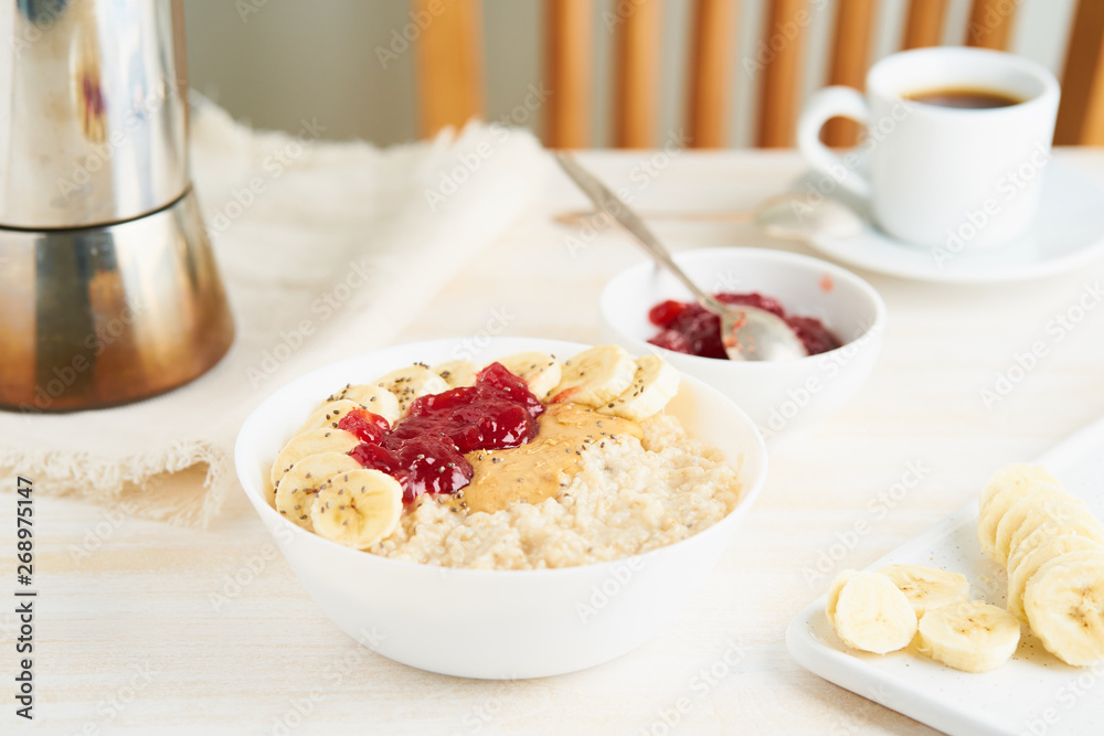 Oatmeal porridge, healthy vegan diet breakfast with strawberry jam, peanut butter, banana, chia on white wooden light background. Side view Oatmeal porridge, healthy vegan diet breakfast with strawberry jam, peanut butter, banana, chia on white wooden light background. Side view