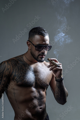 Expensive tobacco. Cigar smoking enjoy life moment. Bearded muscular macho beautiful torso fashion sunglasses smoking cigar. Elite tobacco. Pure enjoyment. Consuming tobacco. Tattooed man with cigar