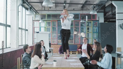 Business woman stands on the table, begins to dance and scatter money and documents. Her colleagues celebrate with her success. Corporate party business team. Trendy office interior. Office life