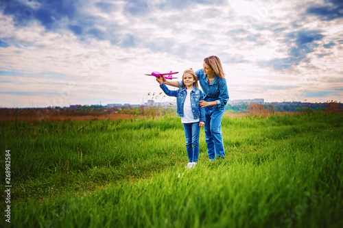 Young woman and girl in denim clothes having fun with small plane on green lawn in summer day