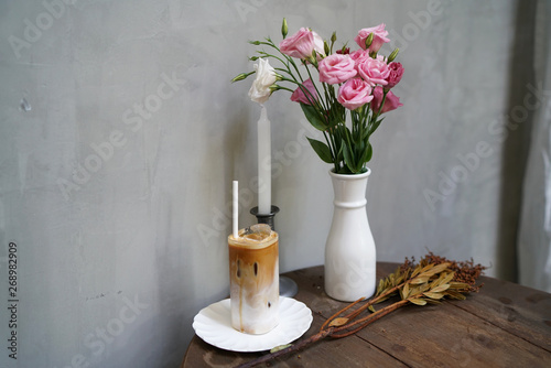 Fototapeta Naklejka Na Ścianę i Meble -  Iced latte - A glass of coffee with milk and vanilla syrup, topped with milk foam on wooden table and flower vase background, Refreshing Summer Drink.