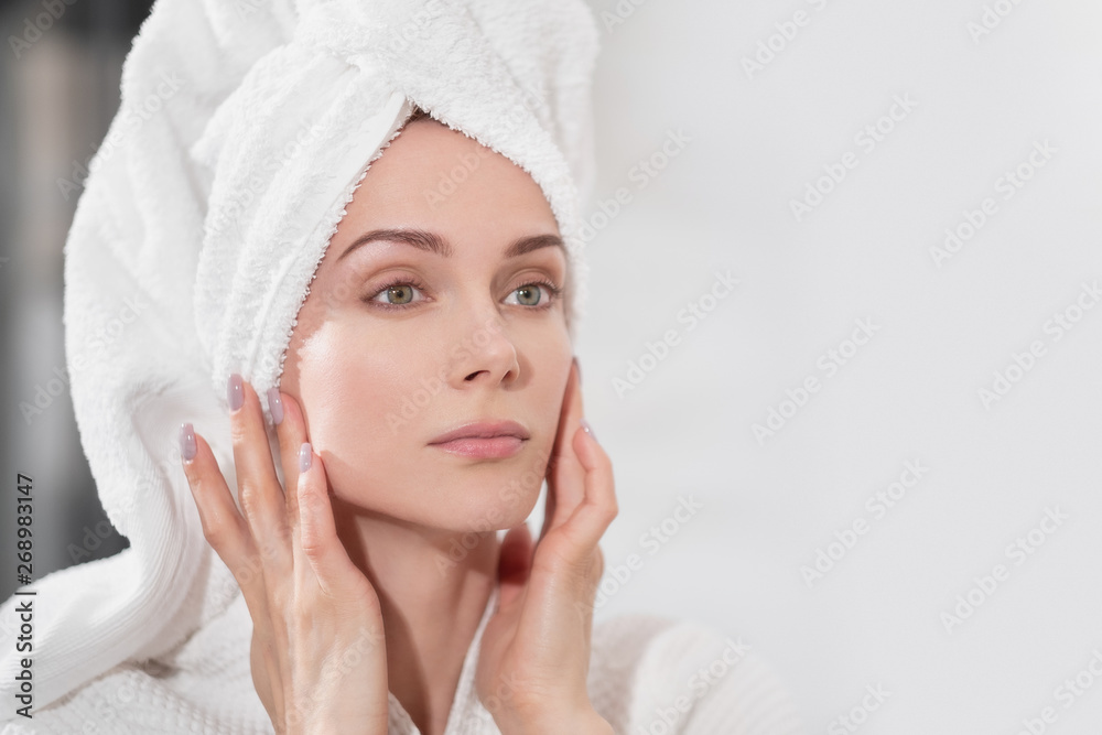 Attractive middle-aged blond woman with white towel on her head and in bathrobe standing in the bathroom by the mirror. She touches skin and smiles. Attractive middle-aged blond woman with white towel on her head and in bathrobe standing in the bathroom by the mirror. She touches skin and smiles.