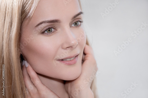 Attractive middle-aged woman a blonde in a white coat stands in the bathroom by the mirror. She touches skin and smiles. Beautiful white teeth lips and eyes.