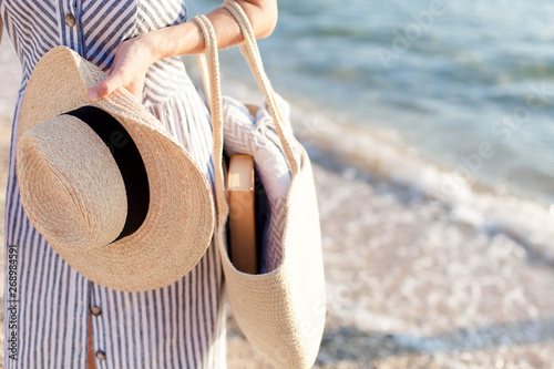 Woman at sea beach. Girl holds straw hat and bag with book and plaid. Traveler is enjoying summer vacation and holidays.