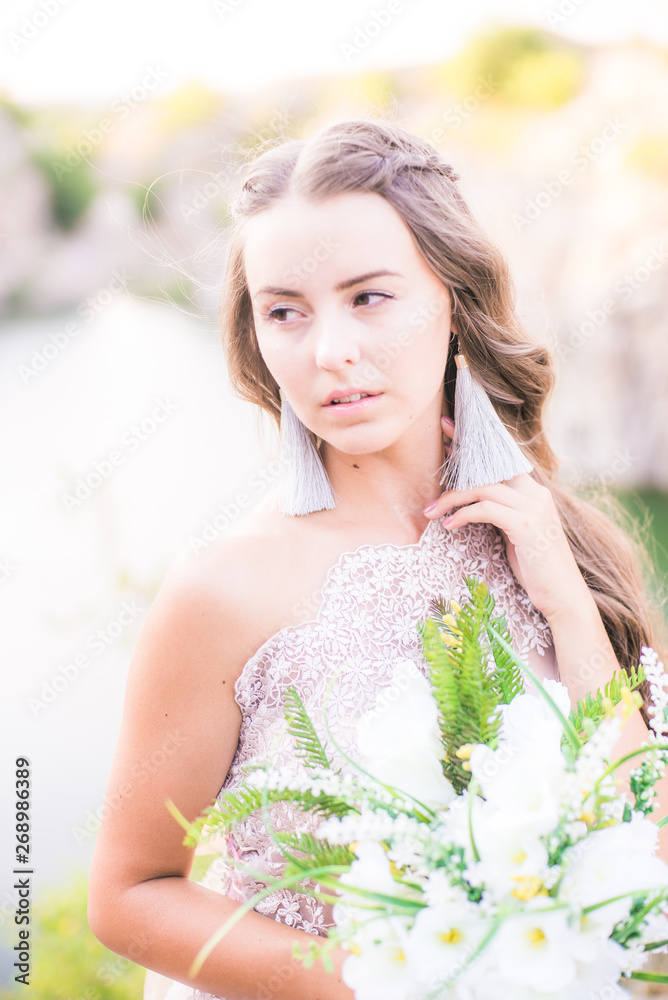 Beautiful young bride with long hair in a wedding dress with a bouquet in the summer on the shore of a beautiful lake Beautiful young bride with long hair in a wedding dress with a bouquet in the summer on the shore of a beautiful lake