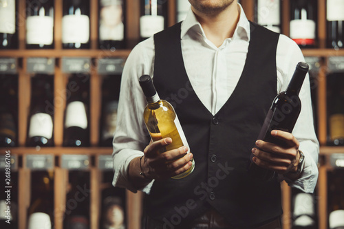 Close-up portrait of sommelier holds a bottle of red and white wine on cellar background. Varieties of alcoholic beverages. photo