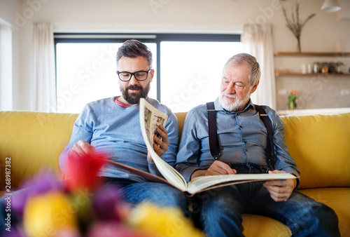 An adult hipster son and senior father sitting on sofa indoors at home.