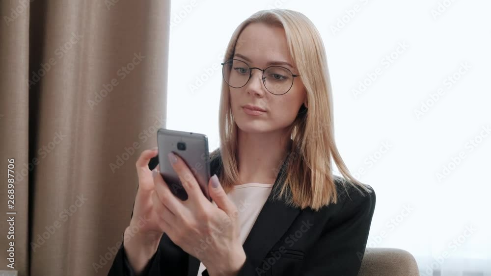 Young attractive middle-aged blonde woman in a black business suit and eyeglasses in a hotel room. Writes messages on the smartphone, works and smiles. Young attractive middle-aged blonde woman in a black business suit and eyeglasses in a hotel room. Writes messages on the smartphone, works and smiles.