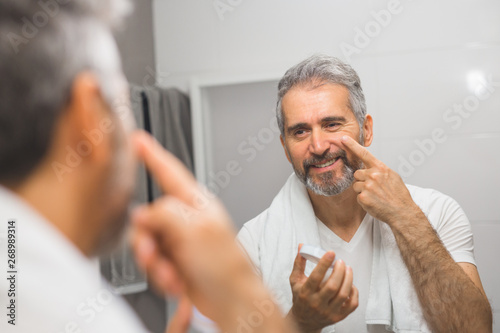 smiling middle aged bearded man applying face cream in bathroom