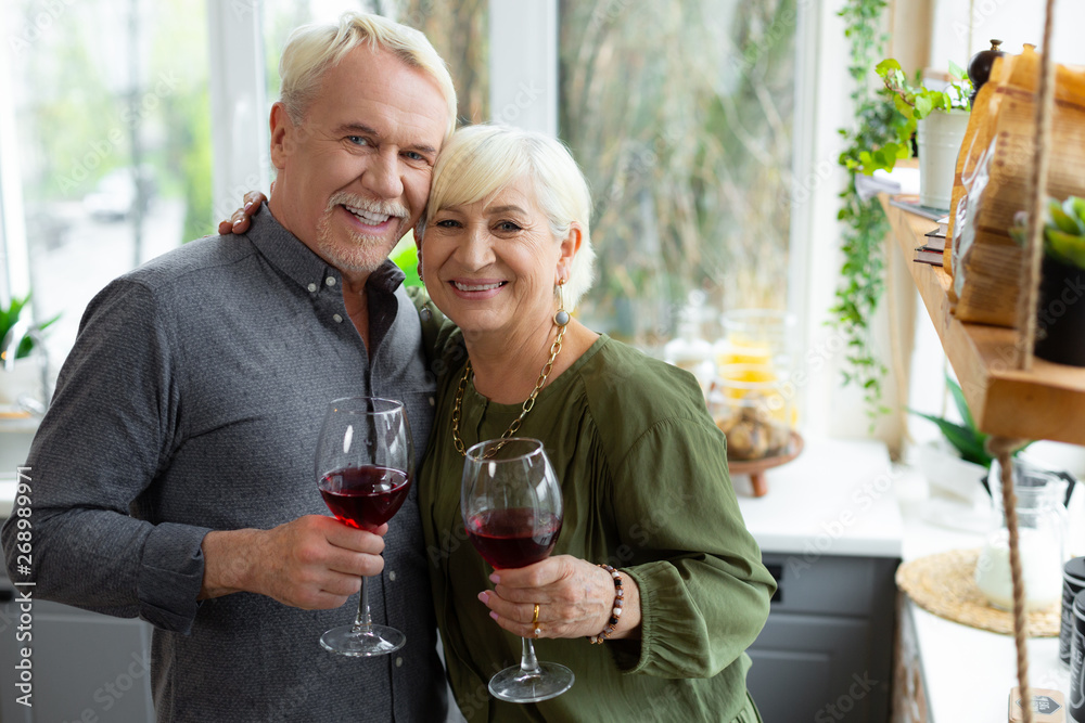 Cheerful silver-haired spouses cuddling and keeping glasses with red wine. Cheerful silver-haired spouses cuddling and keeping glasses with red wine.