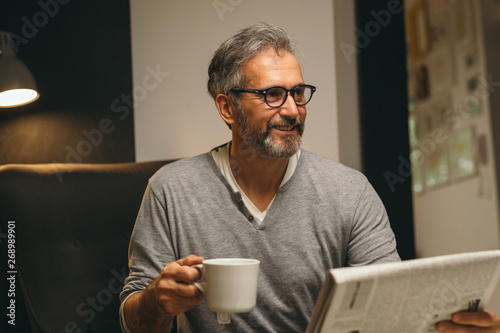 middle aged bearded man reading newspaper and having cup of coffee in his home photo