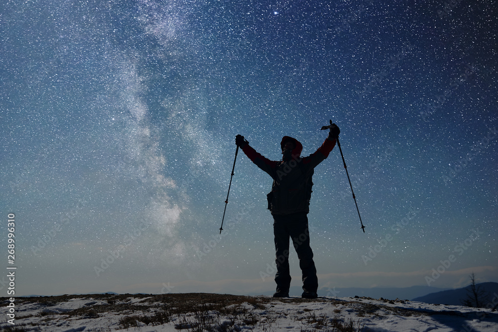 Male silhouette hiker lift his hands up with trekking sticks. Tourist standing on snow-covered top of mountains. Success achievement concept. Starry sky, Milky Way on background. General front view.
