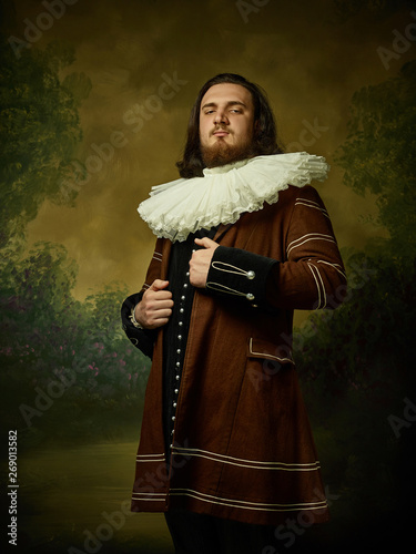 Young man as a medieval knight on dark studio background. Portrait in low key of male model in retro costume. Looking serious. Human emotions, comparison of eras and facial expressions concept.