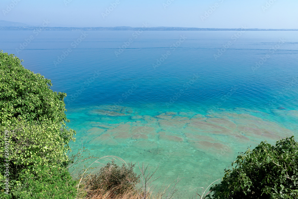 Beautiful emerald and calm waters of Lake Garda near Sirmione town, Italy. On the horizon in the fog the shore with small towns on the lake. Beautiful emerald and calm waters of Lake Garda near Sirmione town, Italy. On the horizon in the fog the shore with small towns on the lake.
