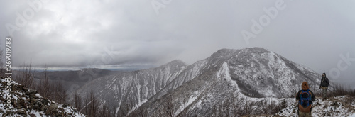 Large panoramic landscape with a ridge  the peaks of which are covered with snow. On the right side of the image  two climbers are visible.