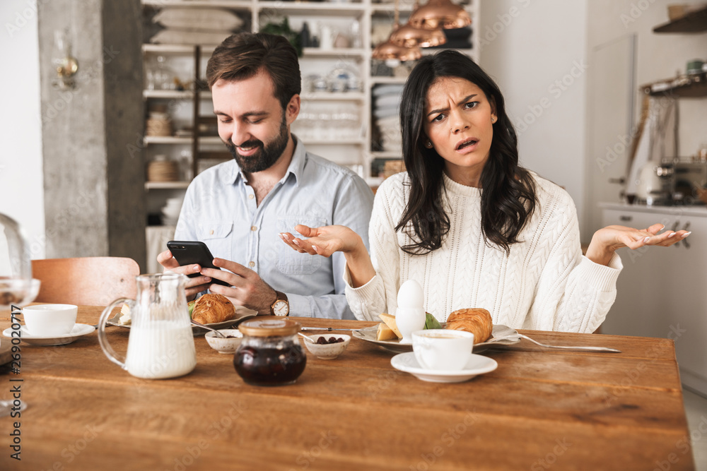 Portrait of european couple using mobile phone while sitting at table together during breakfast in kitchen at home Portrait of european couple using mobile phone while sitting at table together during breakfast in kitchen at home
