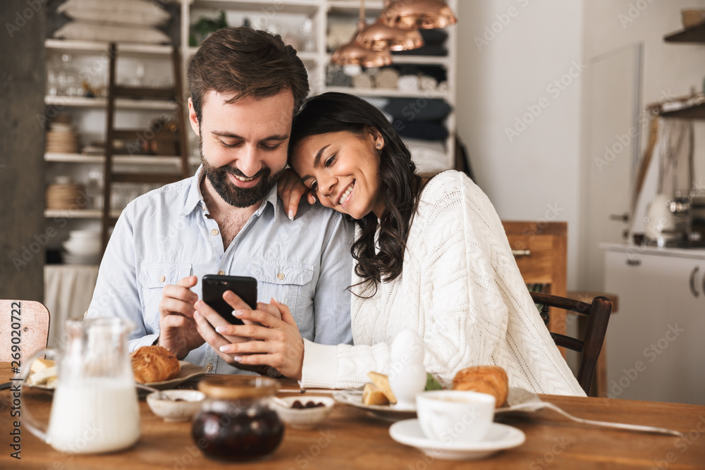 Portrait of happy couple using mobile phone while sitting at table together during breakfast in kitchen at home