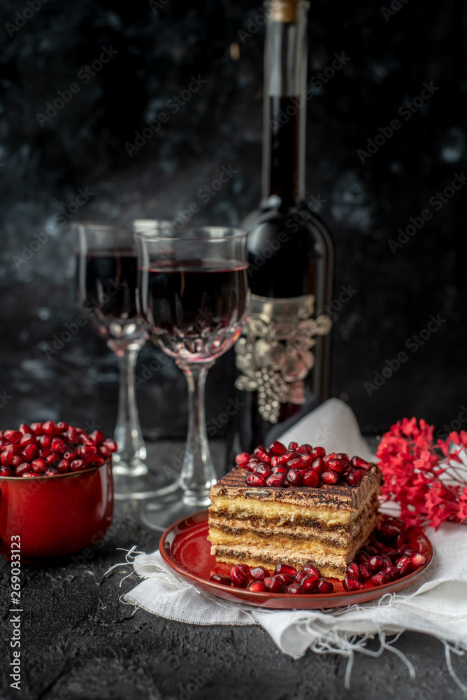 sweet, tempting tiramisu, sprinkled with red grains of pomegranate, on a black stone table, next to it red wine in crystal glasses