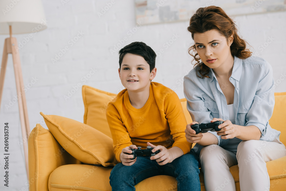 Fototapeta premium focused mother and son playing video game with joysticks while sitting on sofa at home