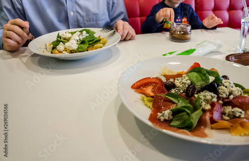 Delicious Italian pasta with vegetables and herbs in a plate on a white table in a restaurant. In the background a man with a child is eating at the table