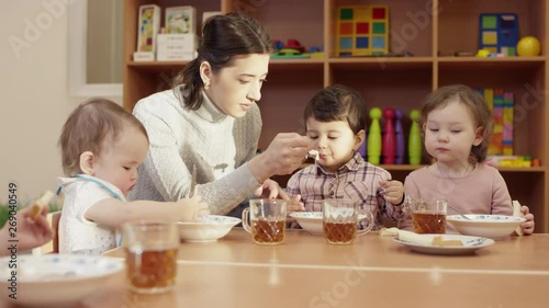 kindergarten teacher sits at the table with the children, and feeds the little dark-haired boy with a spoon, the other children eat on their own, the table is full of plates and mugs, in the photo